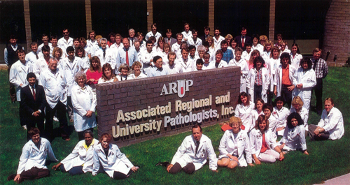 A group of people in lab coats sit on the grass in front of a building
