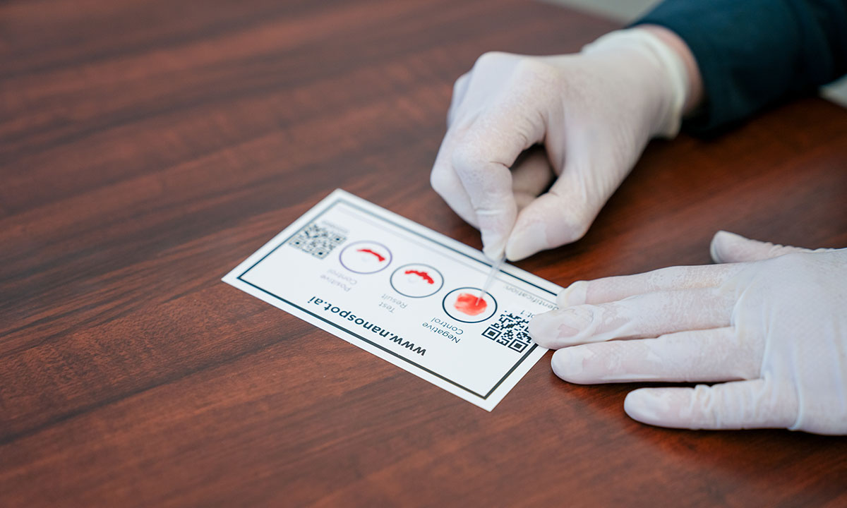 A close-up of two gloved hands. One of the hands is rubbing blood on the NanoSpot.AI rapid COVID-19 antibody test card.