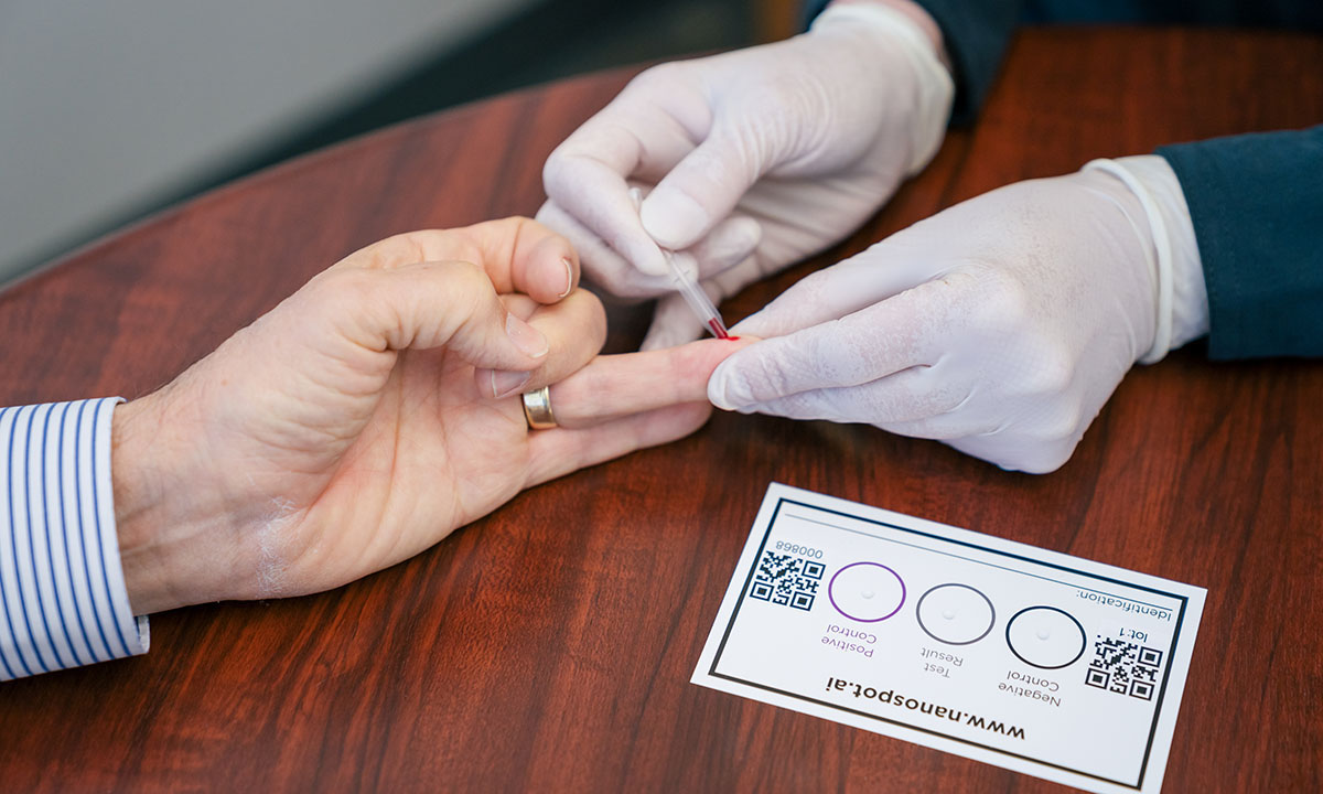 A close-up of three hands. The gloved hands draw blood from a finger of the third hand for the NanoSpot.AI rapid COVID-19 antibody test.