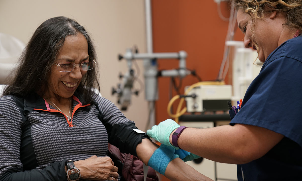 A lab coordinator wraps tape around a sitting woman's arm. They are at the Fourth Street Clinic.