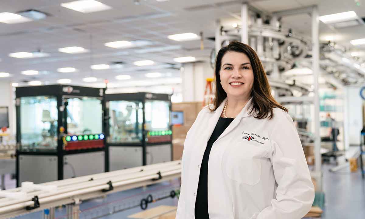 Dr. Tracy George stands in a laboratory. She smiles at the camera.