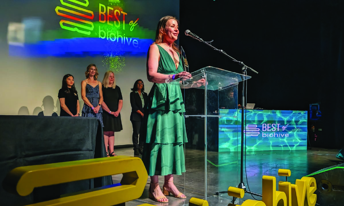 Claire Adam stands in front of a mic. Standing in the background, from left to right, are Kristine Perez, Bonnie Stray, Rachel Lovado, and Janani Sankar.