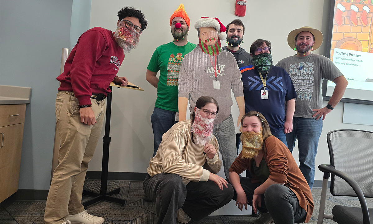 Eight people pose for the camera. They are wearing holiday hats and glitter beards.
