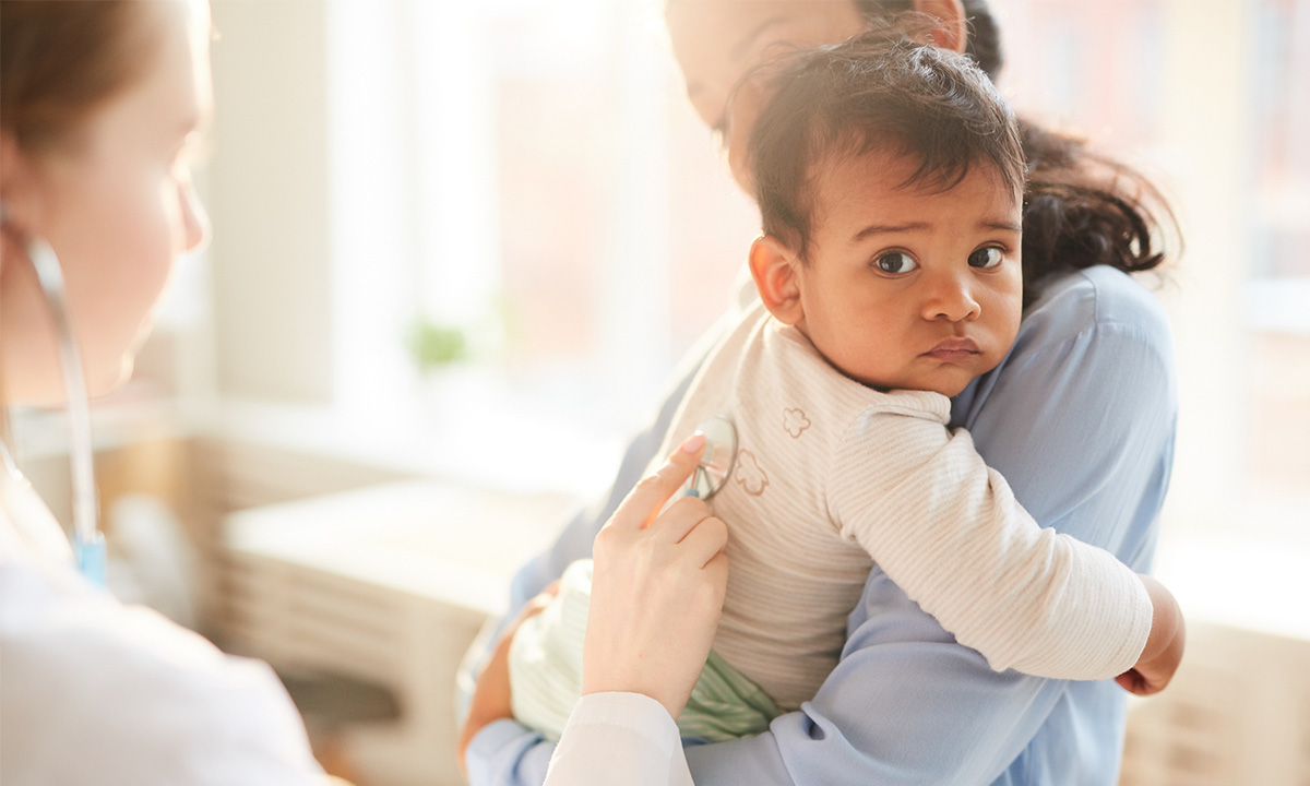 A mother holds her child while a clinician holds a stethoscope on the child's back.