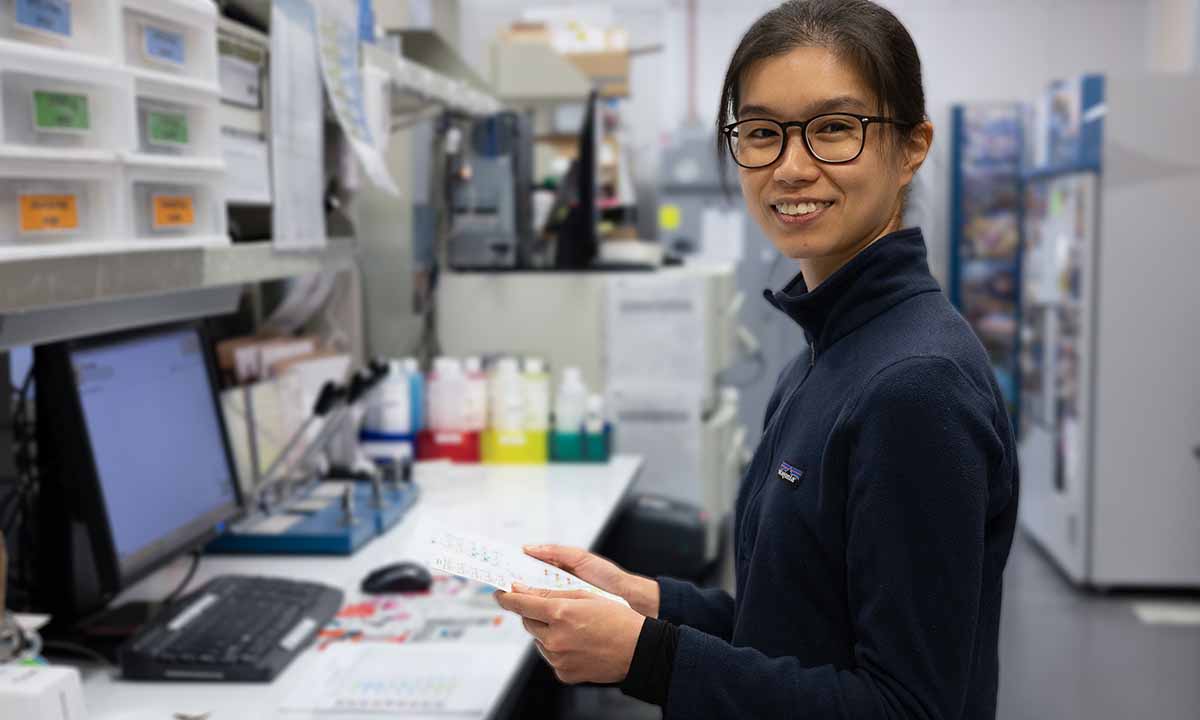 Mei San Tang smiles at the camera while sitting in front of a computer in a lab.