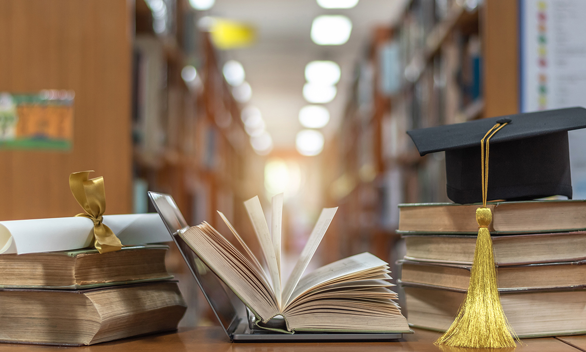 books stacked on a table in a library, with a graduation cap and a rolled-up diploma tied with a ribbon