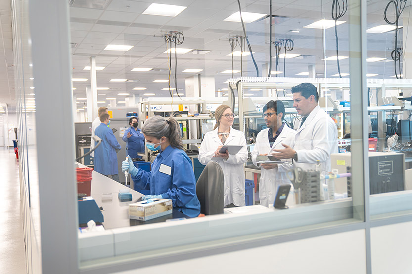 Three people in lab coats watch a lab tech perform her routine