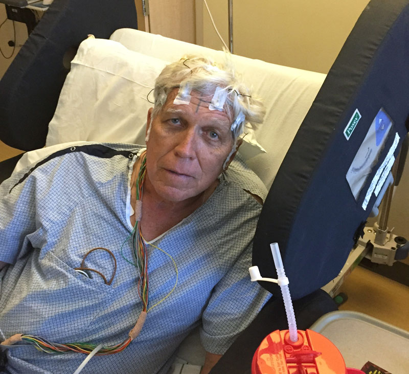 A man in a hospital bed with wires taped to his head