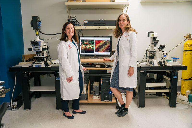 Two women stand in a room with microscopes and computer monitor showing microscopic images