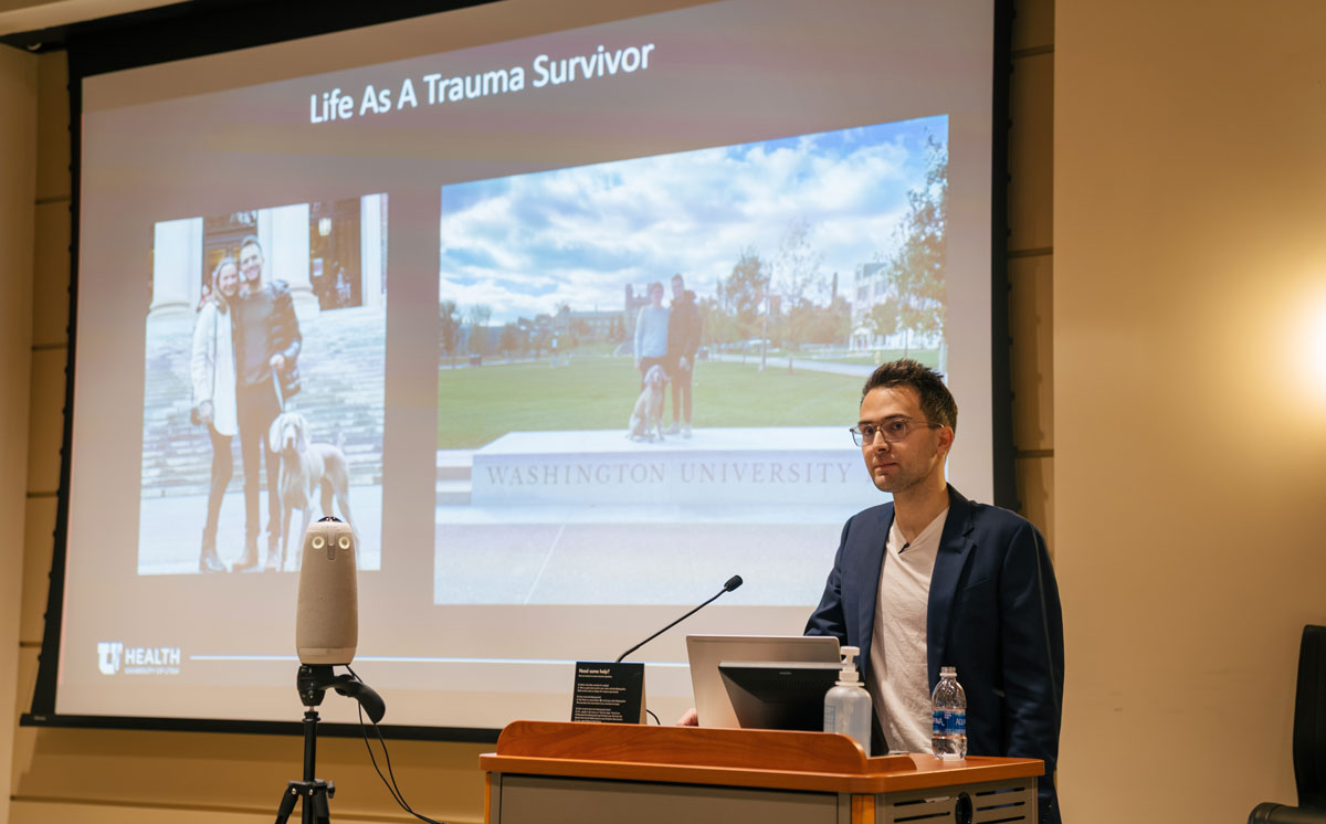 Borges at a podium in front of a screen detailing his life as a trauma survivor